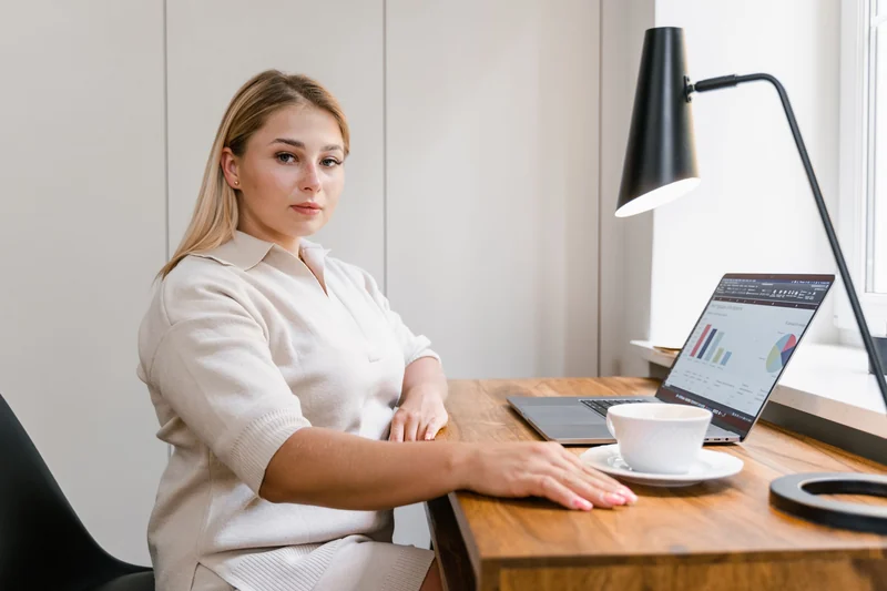 A woman works at a desk by the window, laptop and coffee in front of her.