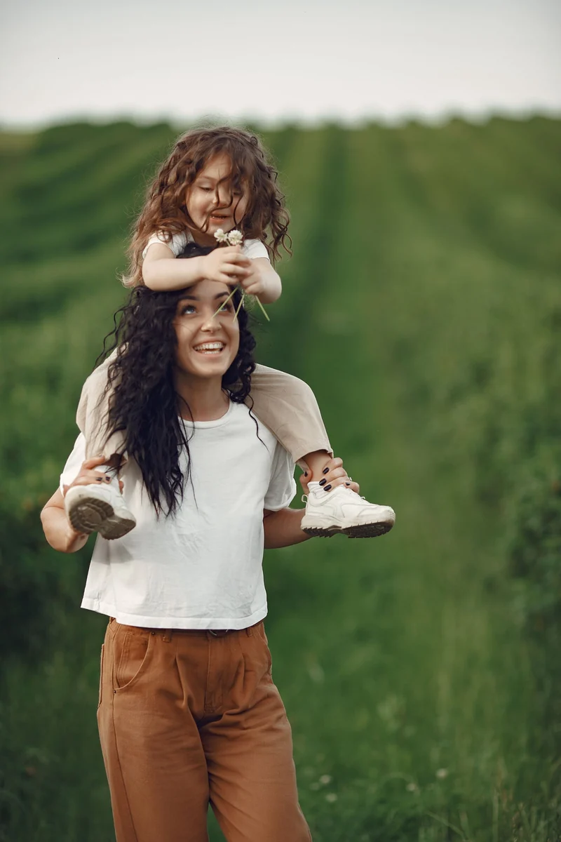 A mother carries her laughing daughter on her shoulders across a meadow.