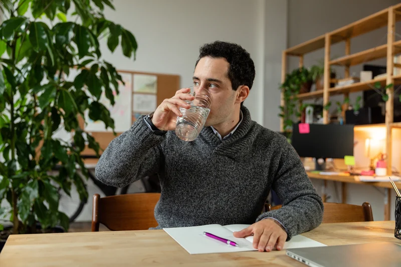 A man in a green knit polo pauses at a table with a glass of water.