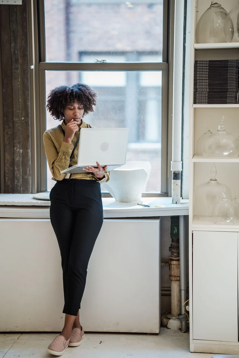 A woman stands by an office window reading on her laptop.