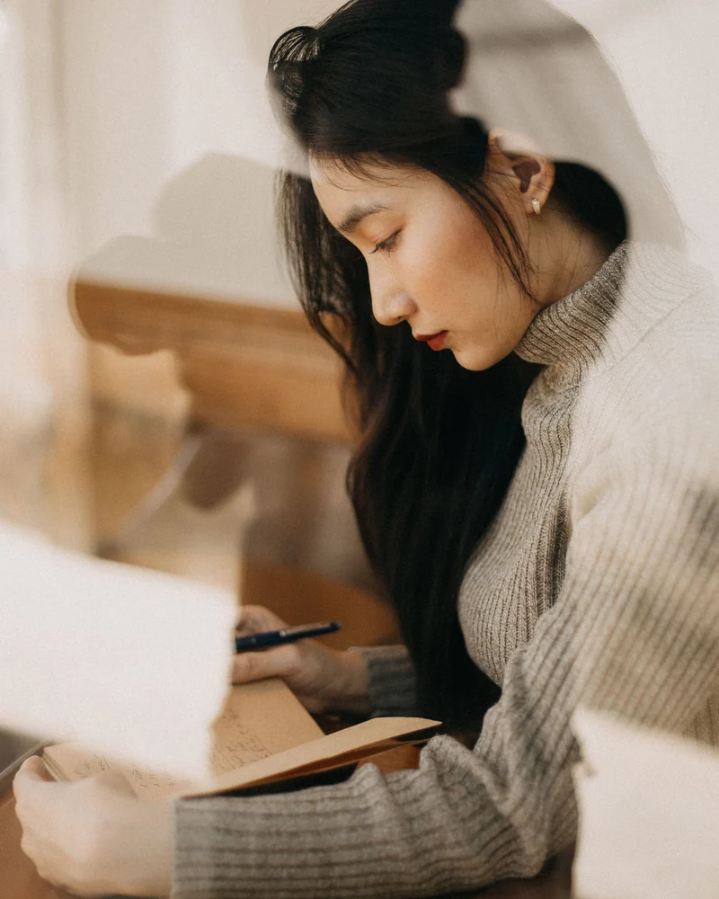 A woman writes in her journal by the window.