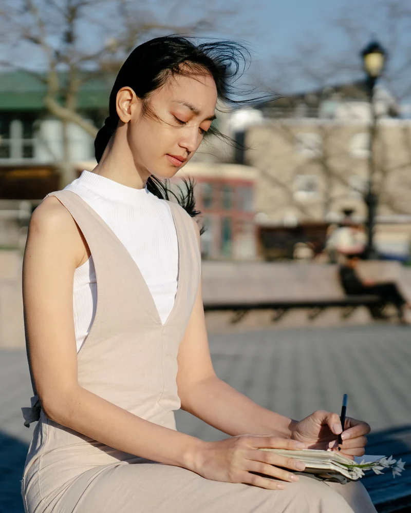 A woman writes in her notebook on a bench in the morning sun.