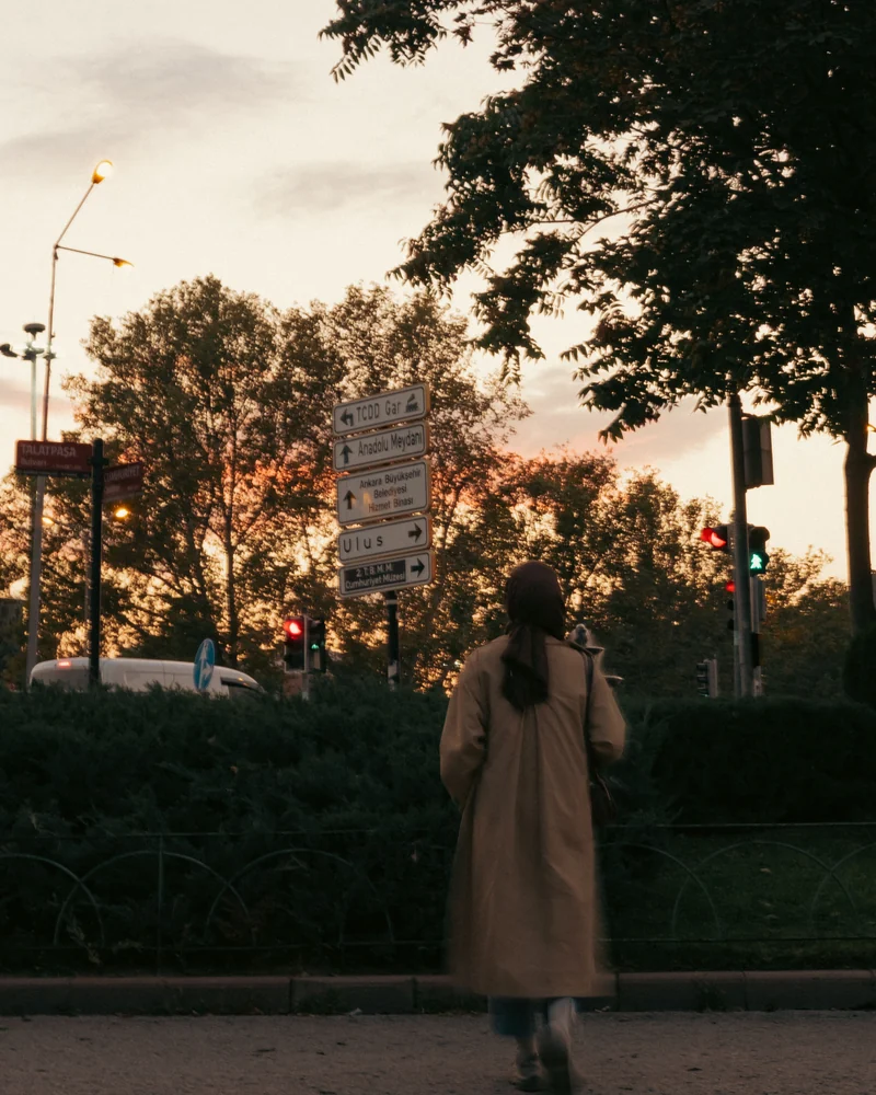 A woman in a trench coat crosses the street at dusk.
