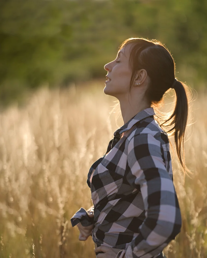 A woman stands in tall grass with her eyes closed, breathing out.