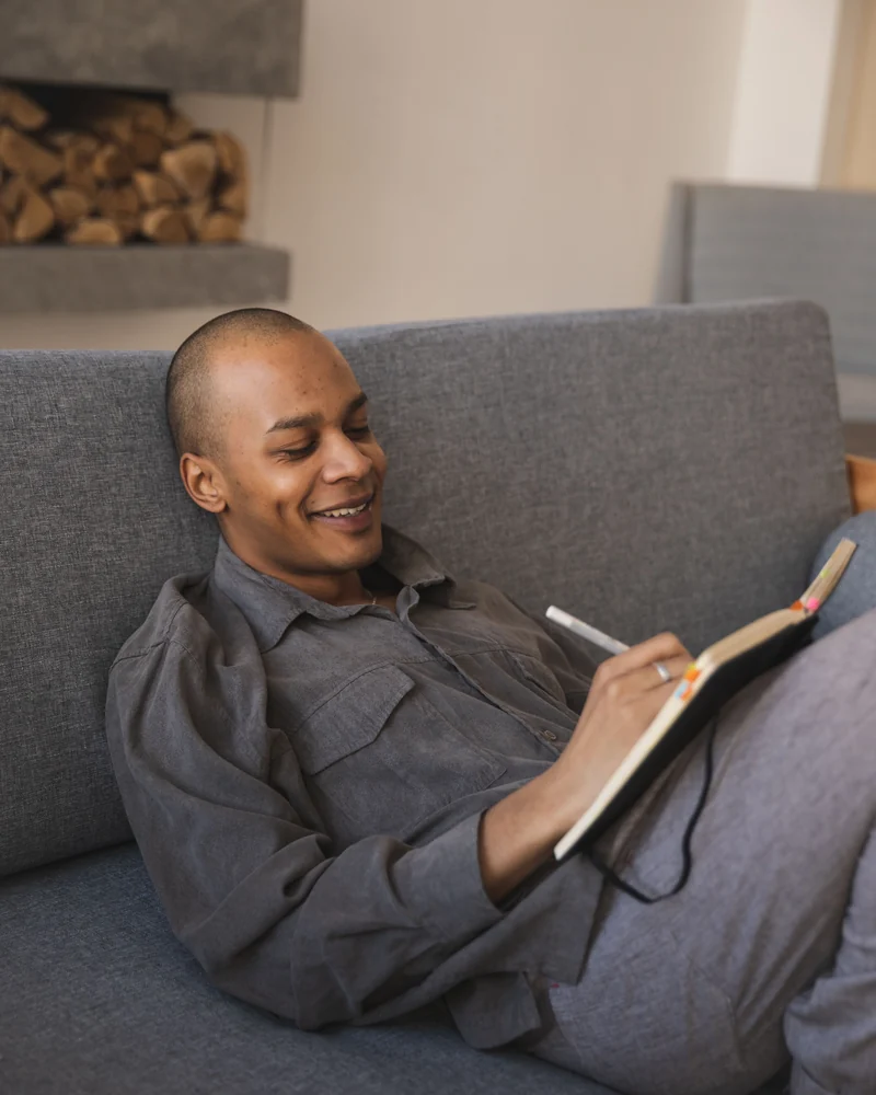 A man smiles on the sofa as he writes in his notebook.